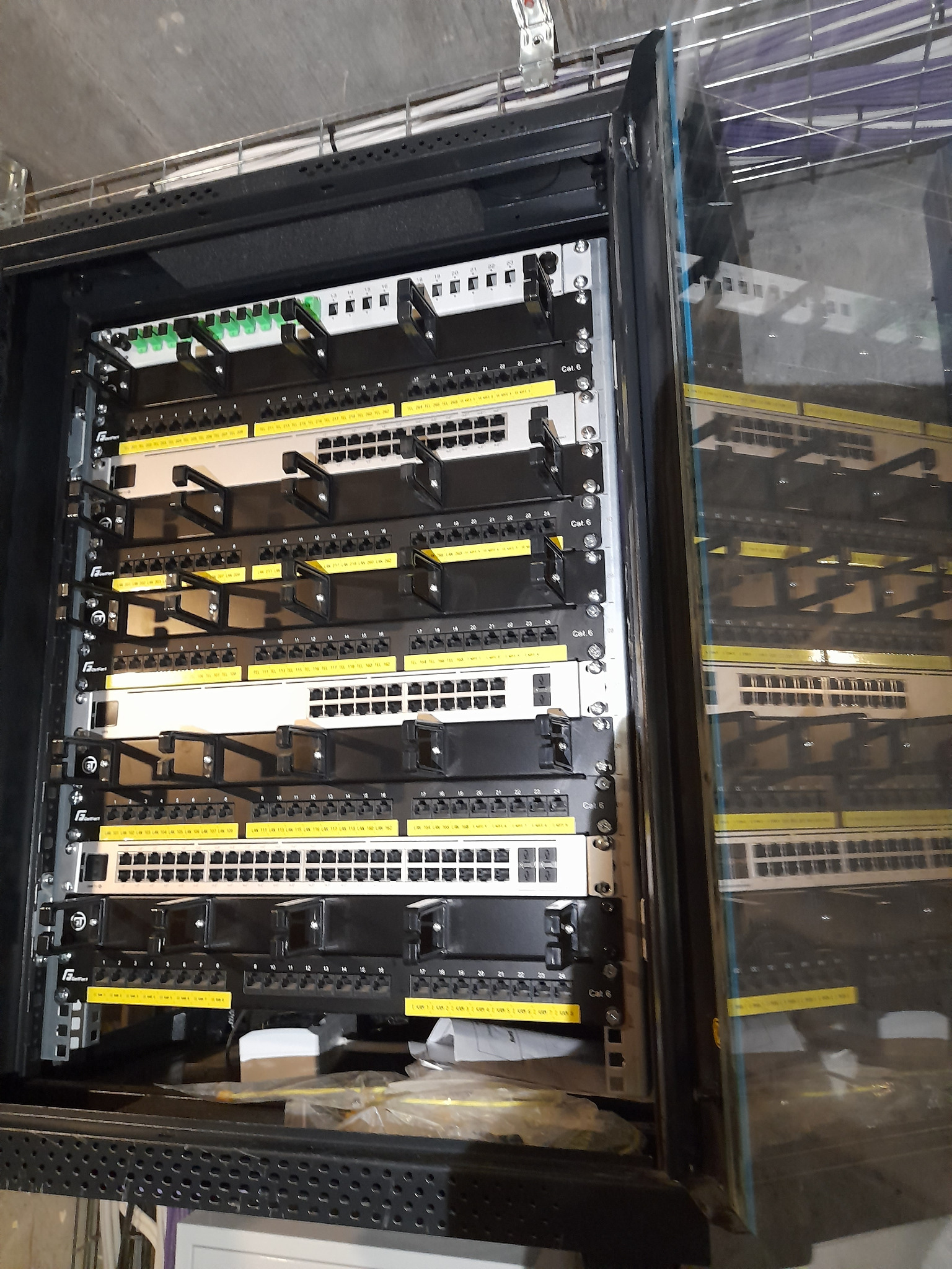 Zamknięta szafa rack z patch panelem Cat 6, widoczne porty ethernet z numeracją i uchwyty kablowe, odblask w szklanych drzwiach szafy.