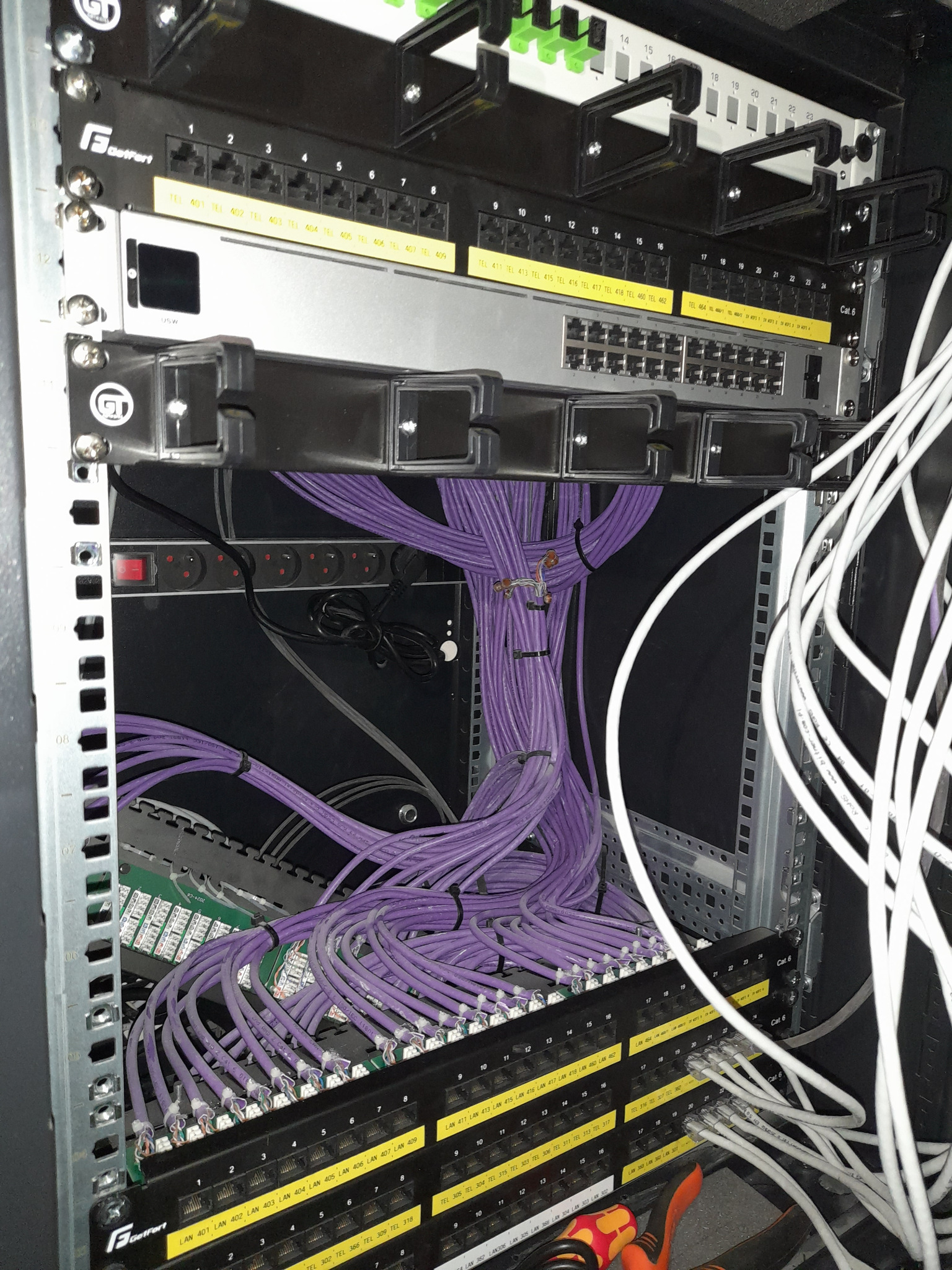 Szafa rack z uporządkowanymi fioletowymi kablami sieciowymi i panelami krosowniczymi, widoczne także białe kable oraz listwa zasilająca.