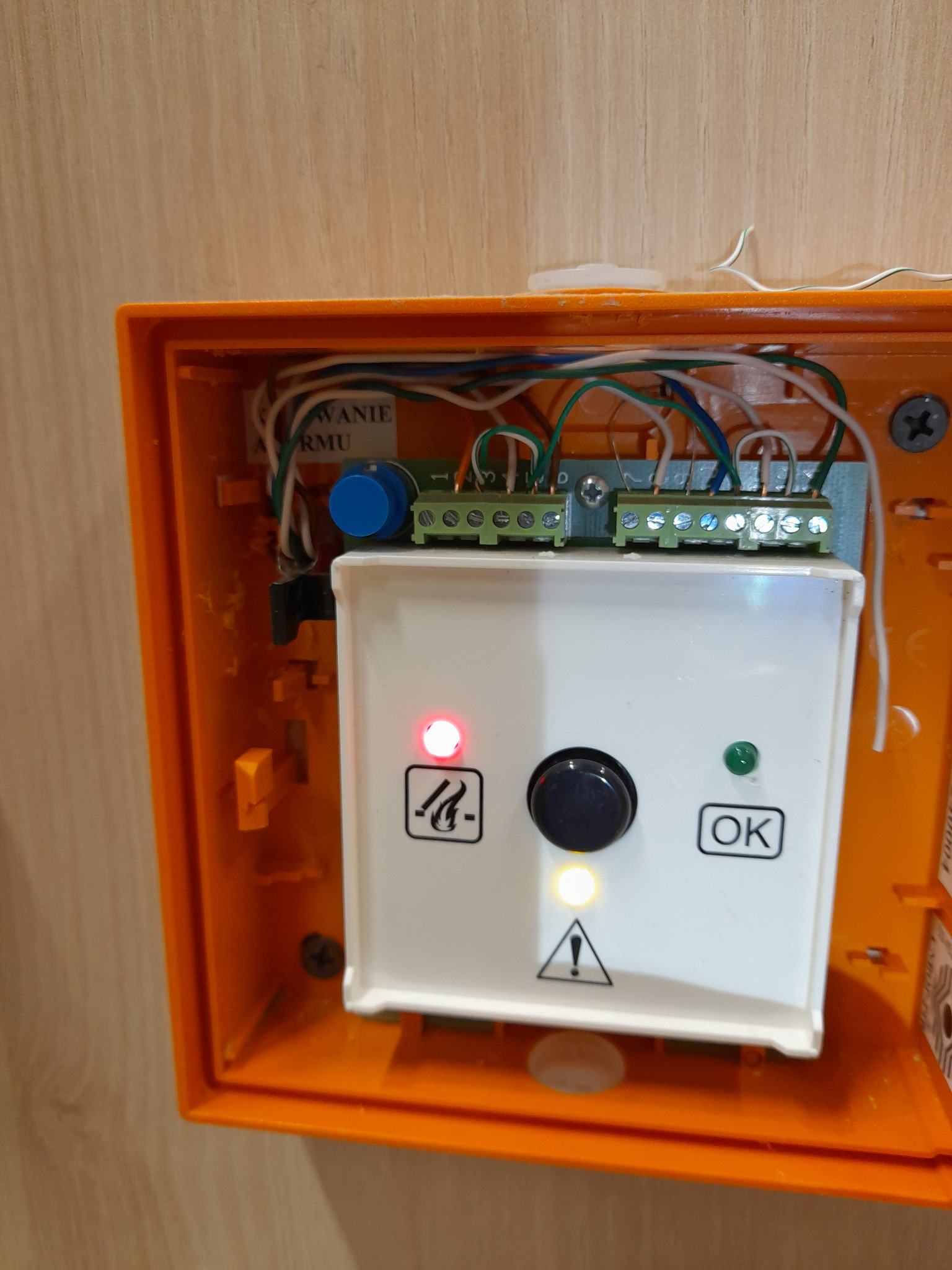 Pomarańczowa skrzynka z białym panelem sterowania alarmem przeciwpożarowym, widoczne diody sygnalizacyjne, przyciski i okablowanie wewnątrz obudowy.