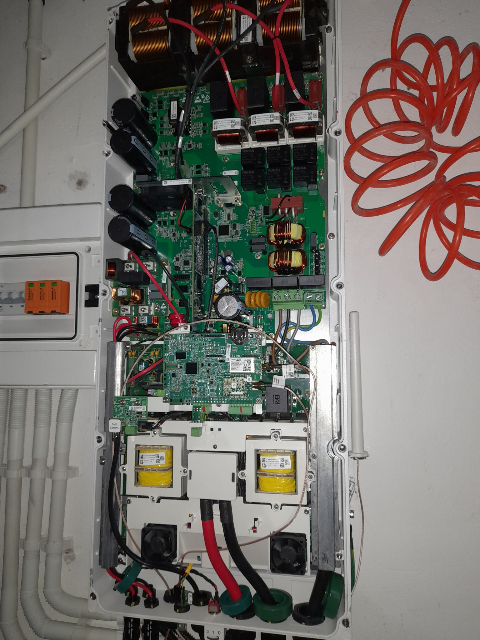 Otwarta obudowa falownika solarnego z widocznymi komponentami elektronicznymi, w tym kondensatorami, transformatorami i płytkami drukowanymi, zamontowana na białej ścianie obok rur instalacyjnych...