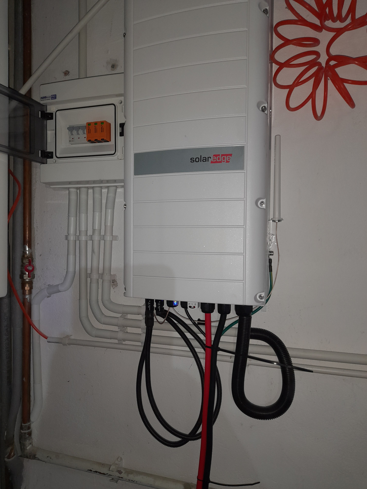 Szara skrzynka SolarEdge zamontowana na ścianie obok otwartej skrzynki z bezpiecznikami i rurkami instalacyjnymi.