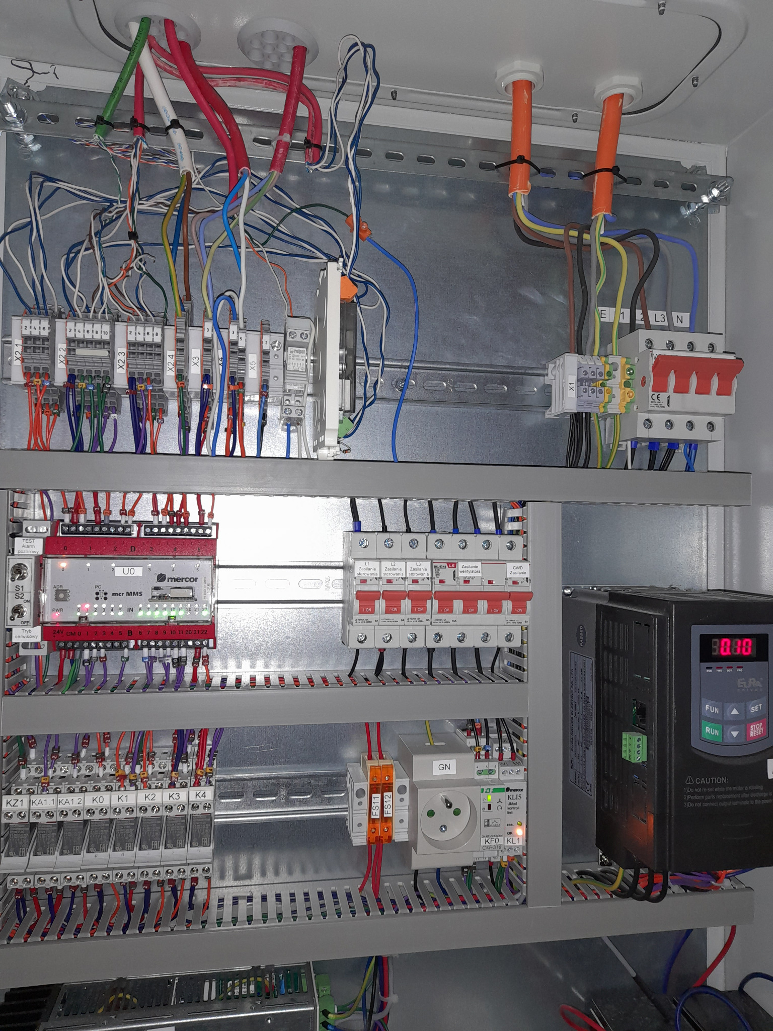 Wnętrze szafy sterowniczej z licznymi przewodami, modułami, zabezpieczeniami i sterownikiem silnika z wyświetlaczem LED. Widoczne kable w różnych kolorach, listwy zaciskowe, przekaźniki oraz zasilacz.