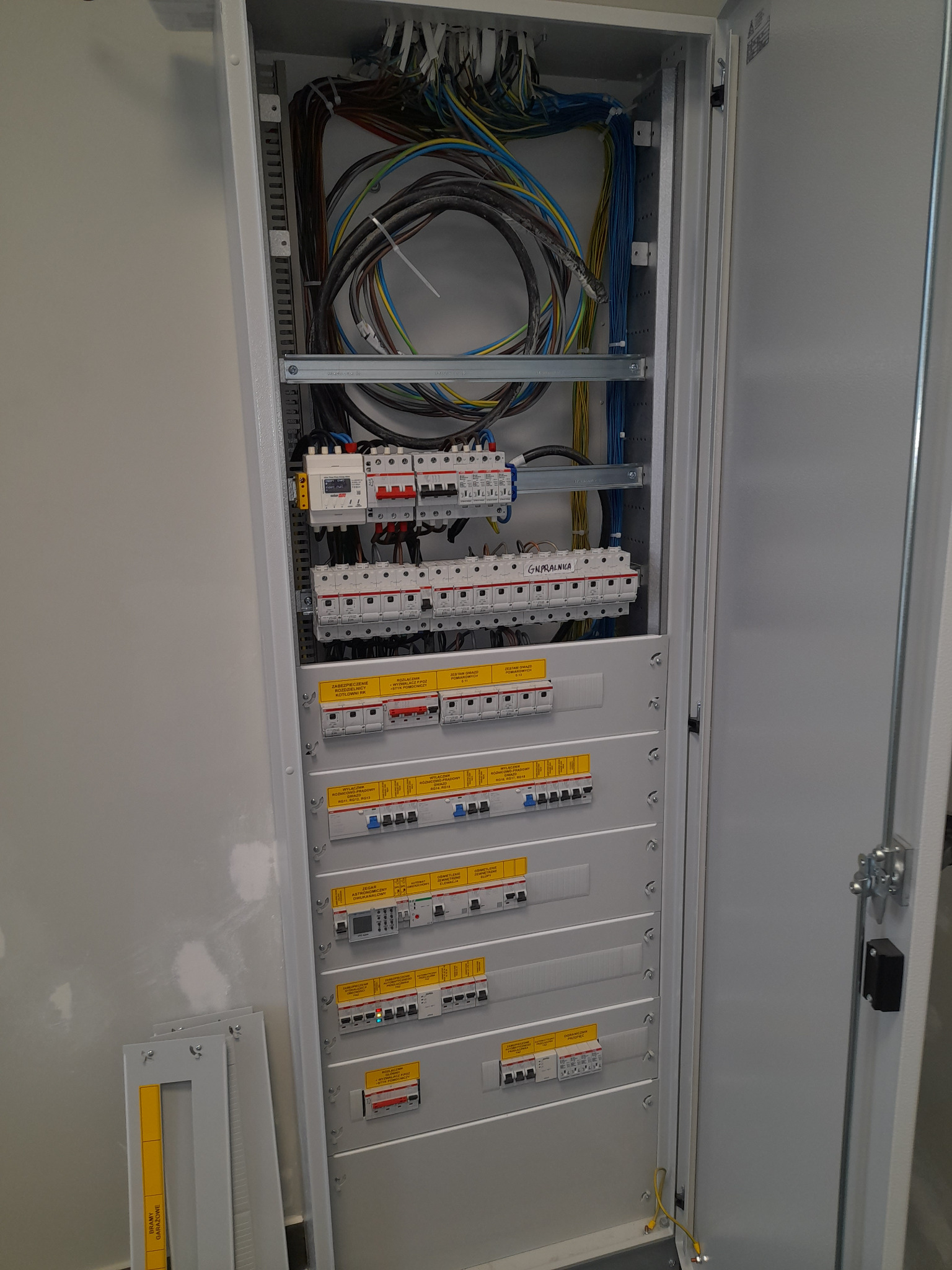 Otwarta szafa rozdzielcza z widocznymi zabezpieczeniami, przewodami i elementami montażowymi. Na dole leżą elementy do montażu. Widoczne żółte etykiety na bezpiecznikach.