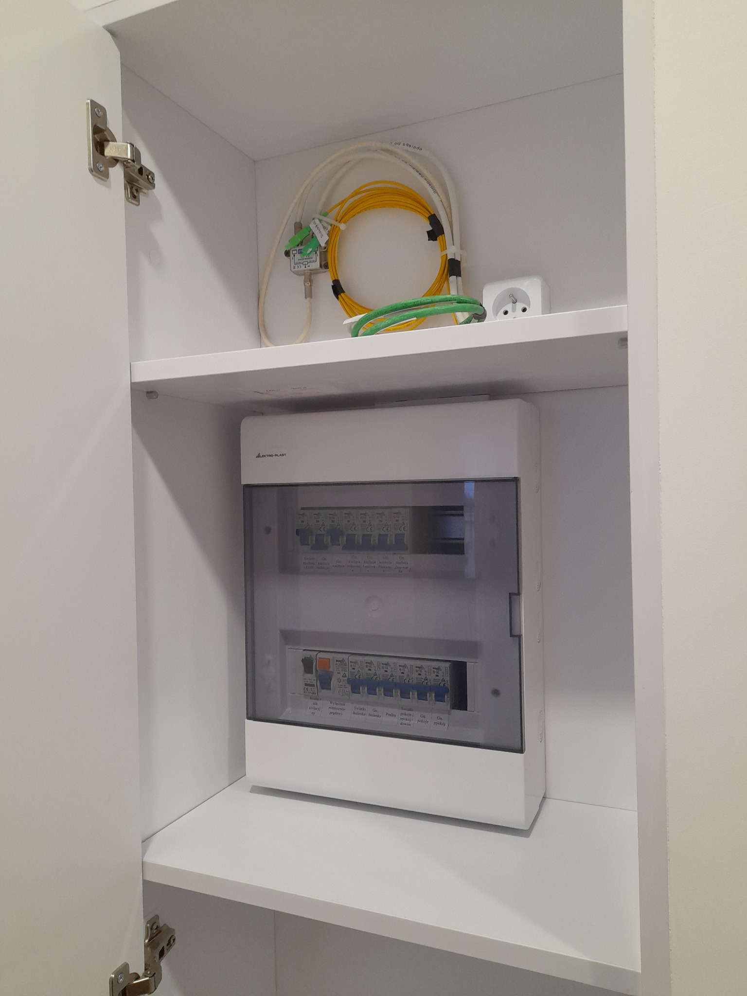 Biała szafka z rozdzielnicą elektryczną Elektro-Plast, widoczne kable światłowodowe i gniazdko elektryczne na górnej półce.