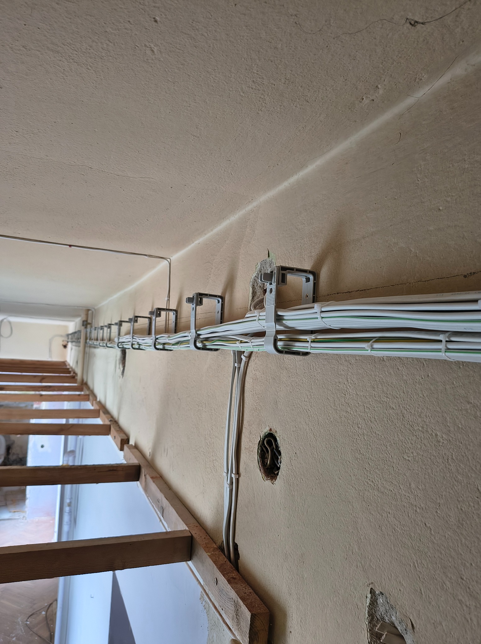 Białe kable elektryczne prowadzone wzdłuż ściany, zamocowane za pomocą uchwytów. Widoczne otwory w ścianie i drewniana konstrukcja. Ujęcie z dołu.