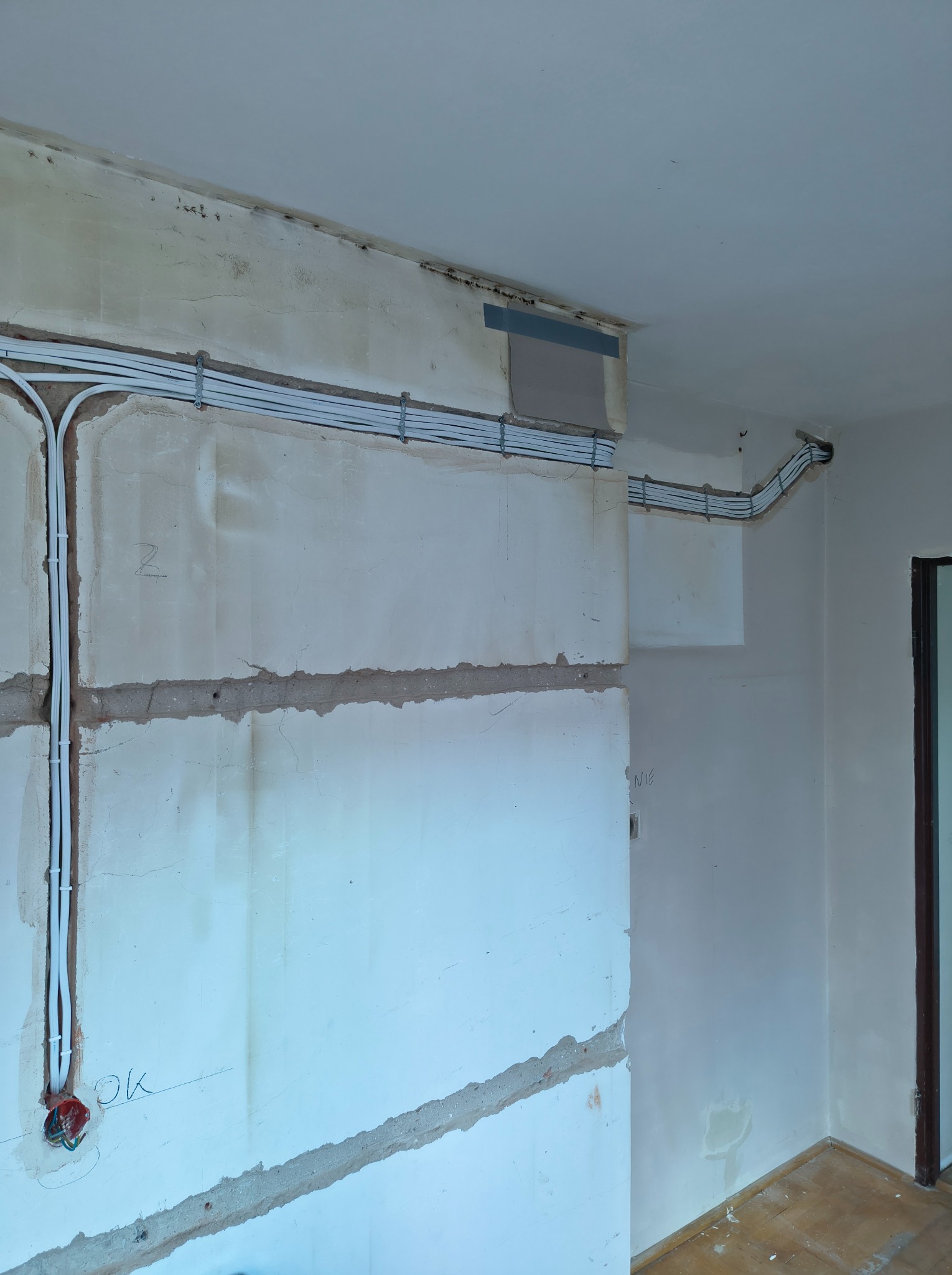 Białe kable elektryczne ułożone wzdłuż surowej ściany w trakcie remontu. Widoczne uchwyty mocujące i puszka instalacyjna. Fragment podłogi i drzwi w tle.