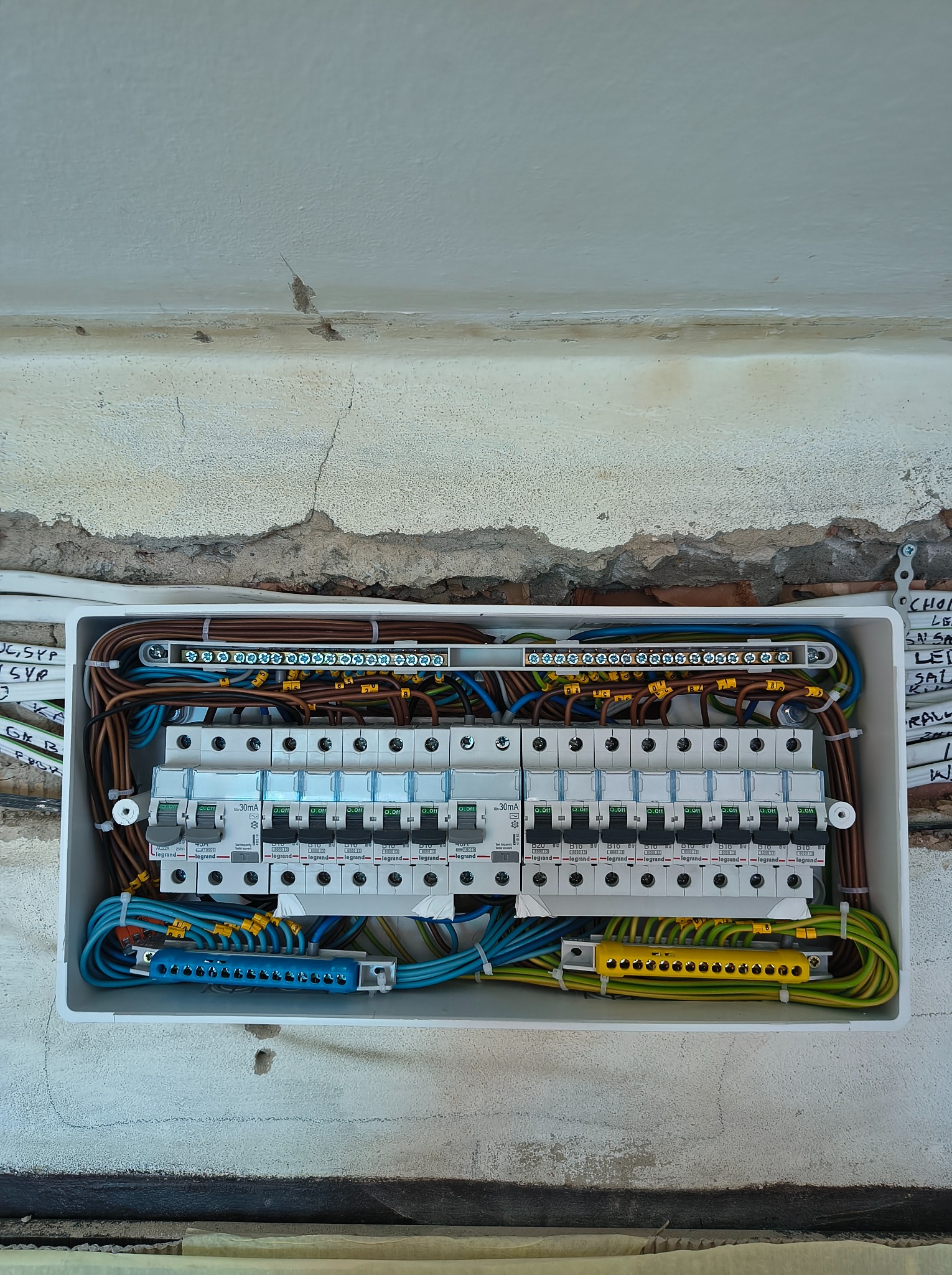 Otwarta skrzynka rozdzielcza z bezpiecznikami Legrand, widoczne kable w różnych kolorach, zamontowana na nierównej ścianie z widocznymi śladami po instalacji.