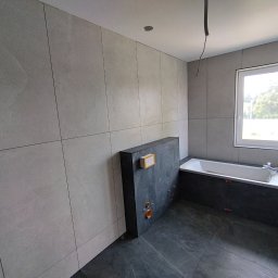 Ołeksij Moroz - Wnętrze łazienki w trakcie wykańczania, widoczne szare płytki na ścianach i podłodze, zabudowa wanny z ciemnego kamienia, przygotowane przyłącza hydrauliczne oraz wystające kable z sufitu.