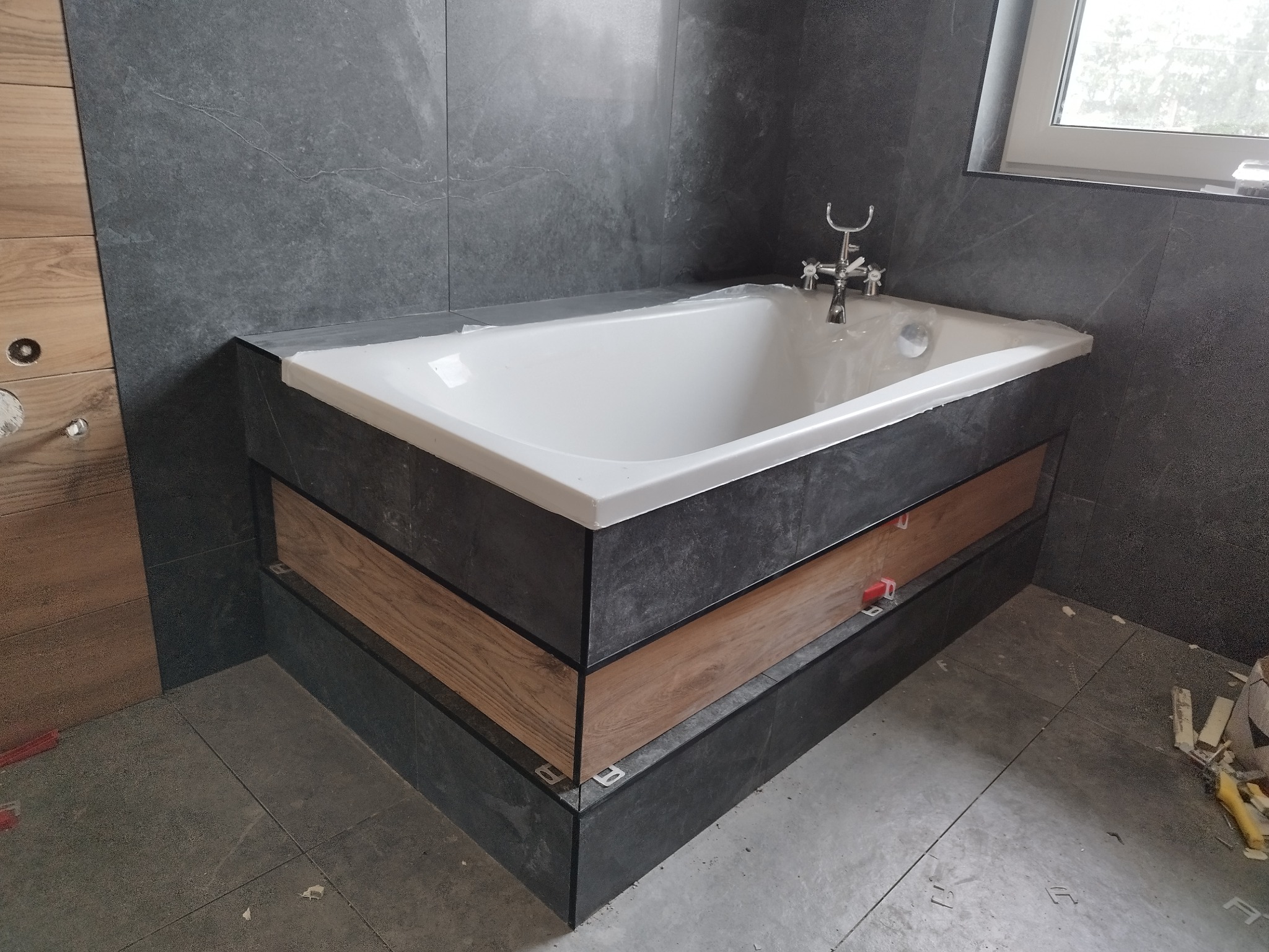 Wanna obudowana szarą płytką i drewnopodobną wstawką, widoczne krzyżyki glazurnicze, szare płytki na ścianach i podłodze, bateria wannowa w stylu retro.