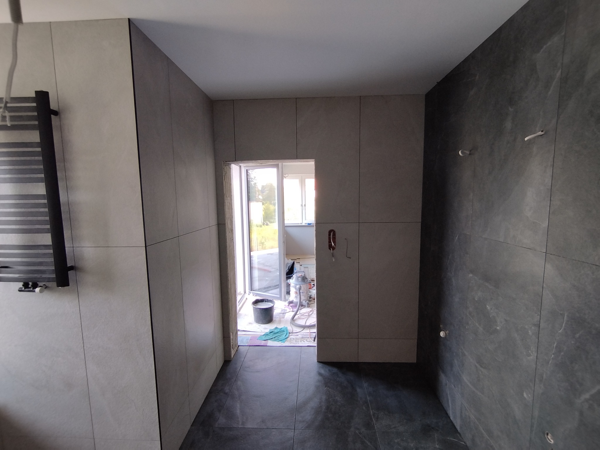 Wnętrze łazienki w trakcie wykańczania: szare płytki na ścianach i podłodze, widoczne narzędzia i materiały budowlane przy wejściu balkonowym, grzejnik drabinkowy po lewej.