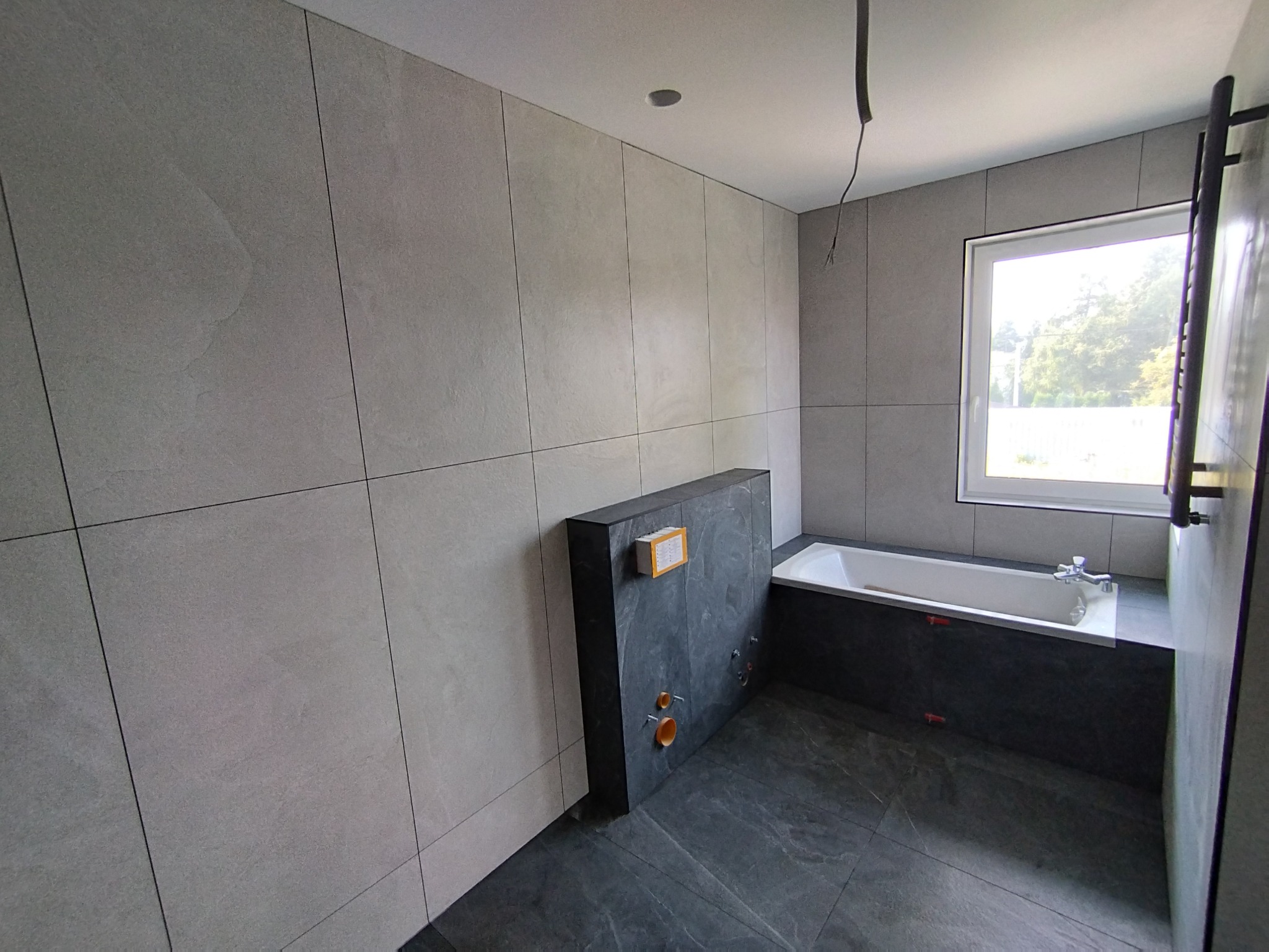 Wnętrze łazienki w trakcie wykańczania, widoczne szare płytki na ścianach i podłodze, zabudowa wanny z ciemnego kamienia, przygotowane przyłącza hydrauliczne oraz wystające kable z sufitu.