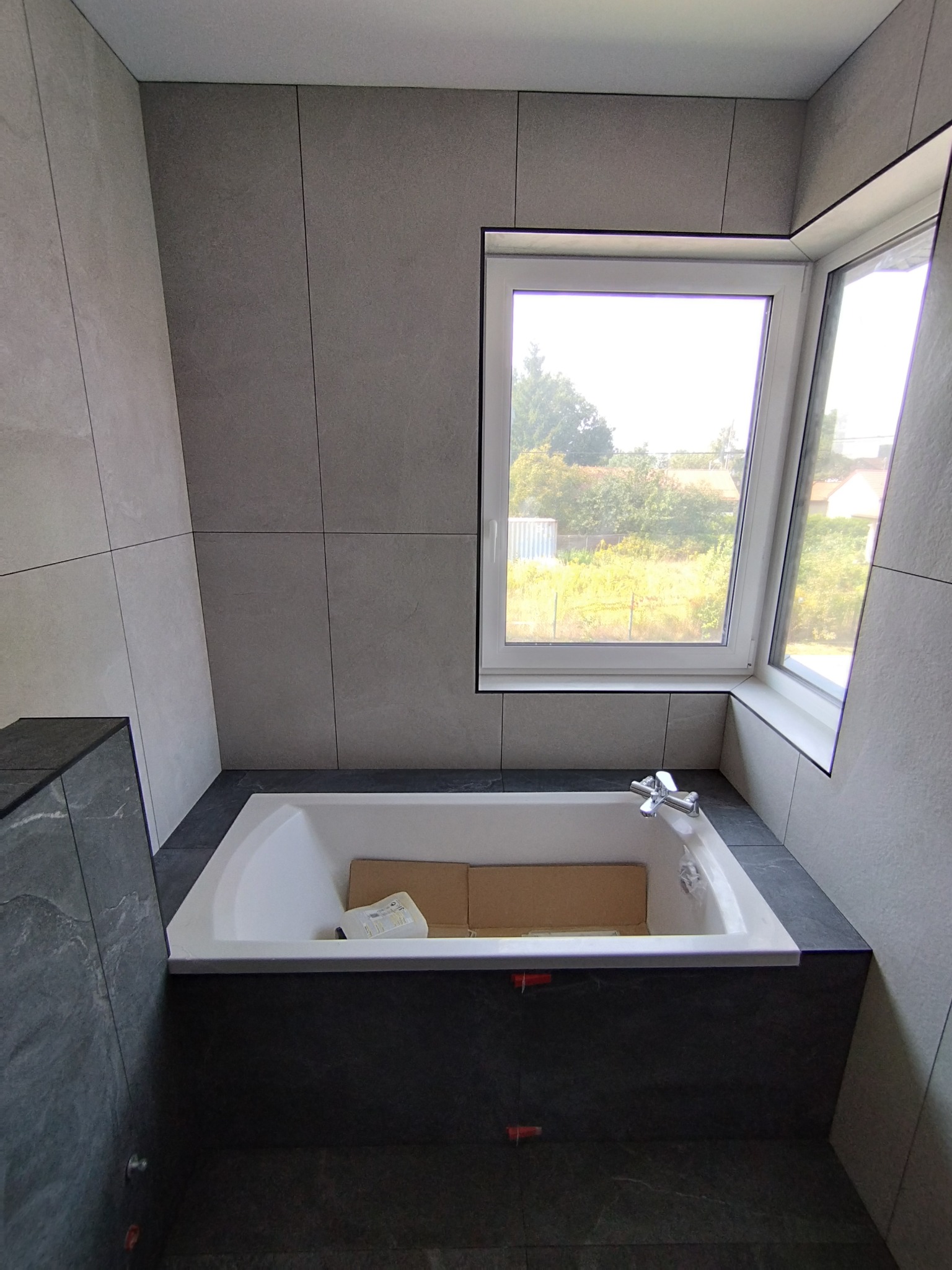 Wnętrze łazienki w trakcie prac wykończeniowych: wanna obudowana płytkami, okno z widokiem na zieleń, szare płytki na ścianach, widoczne krzyżyki dystansowe na obudowie wanny.