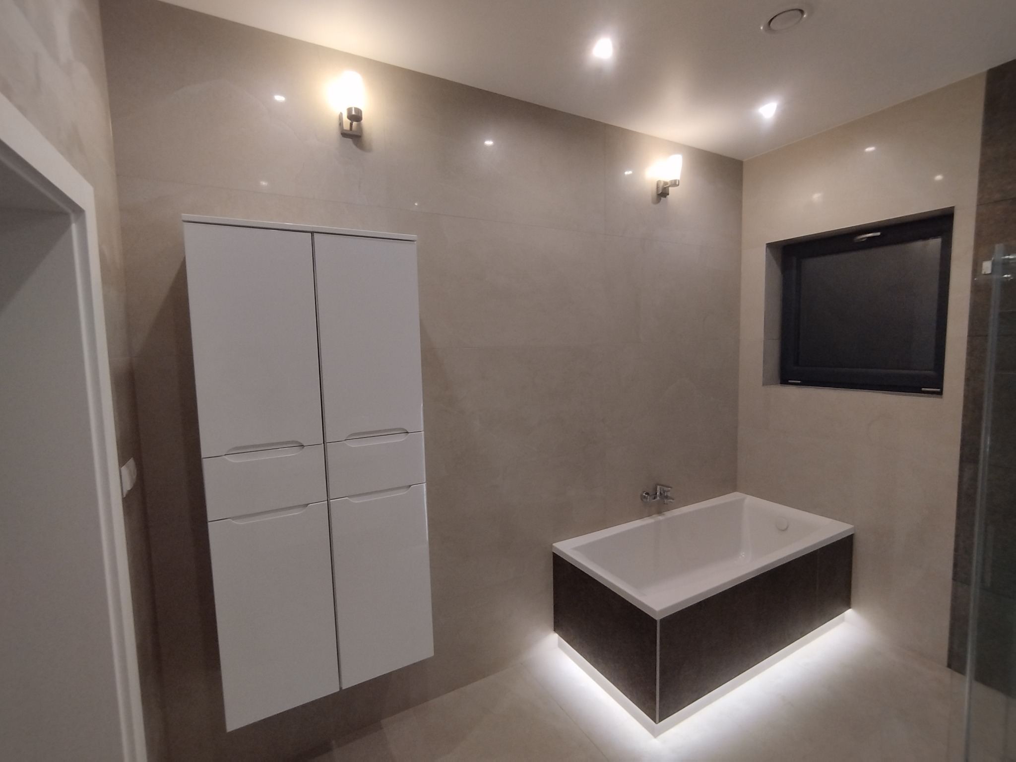 Nowoczesna łazienka z podświetlaną wanną, białą szafką wiszącą i beżowymi płytkami ściennymi, oświetlona kinkietami i punktowym światłem sufitowym.