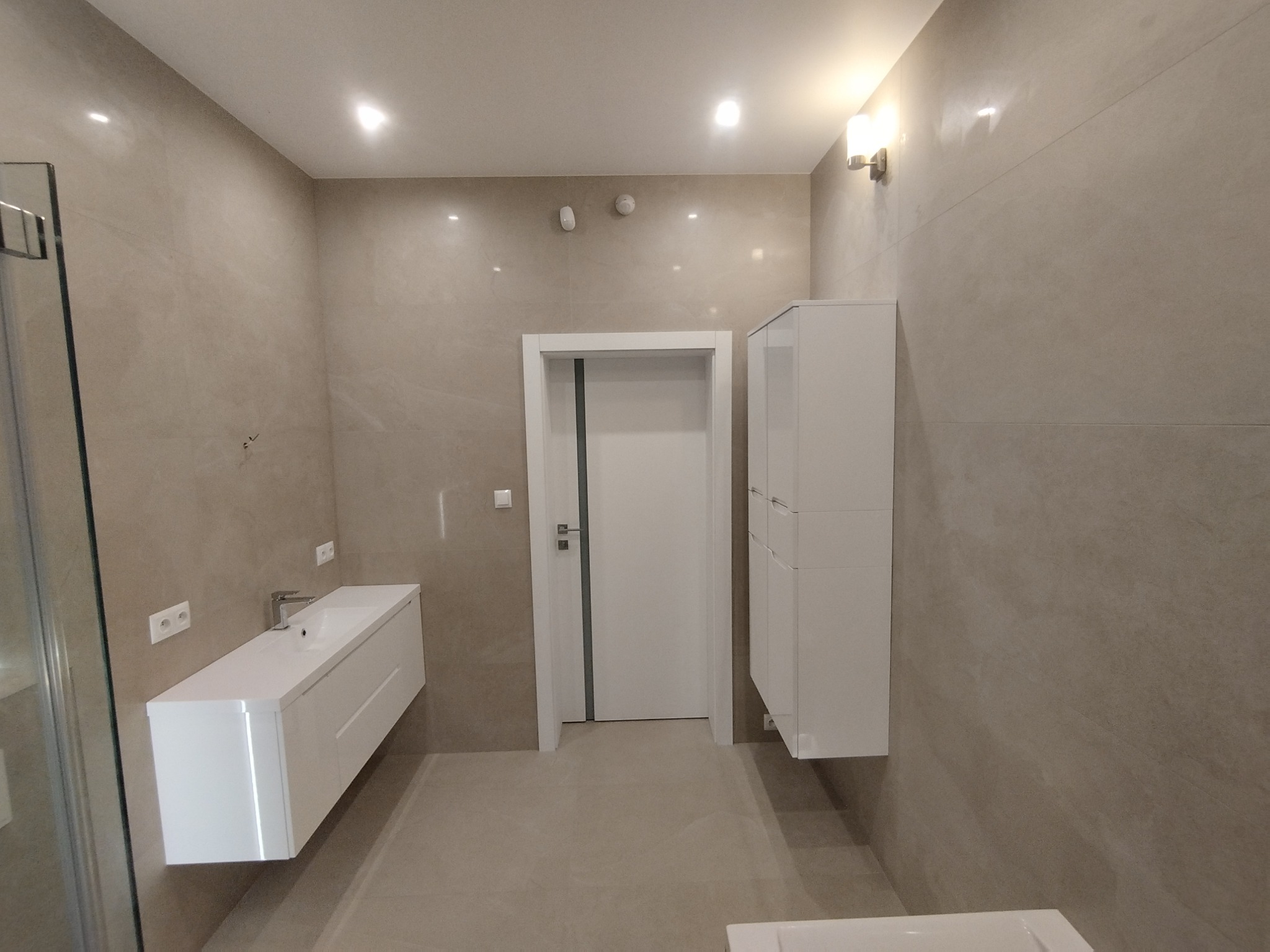 Nowoczesna łazienka z beżowymi płytkami, białą szafką pod umywalką, szafką wiszącą i drzwiami, oświetlona kinkietem.