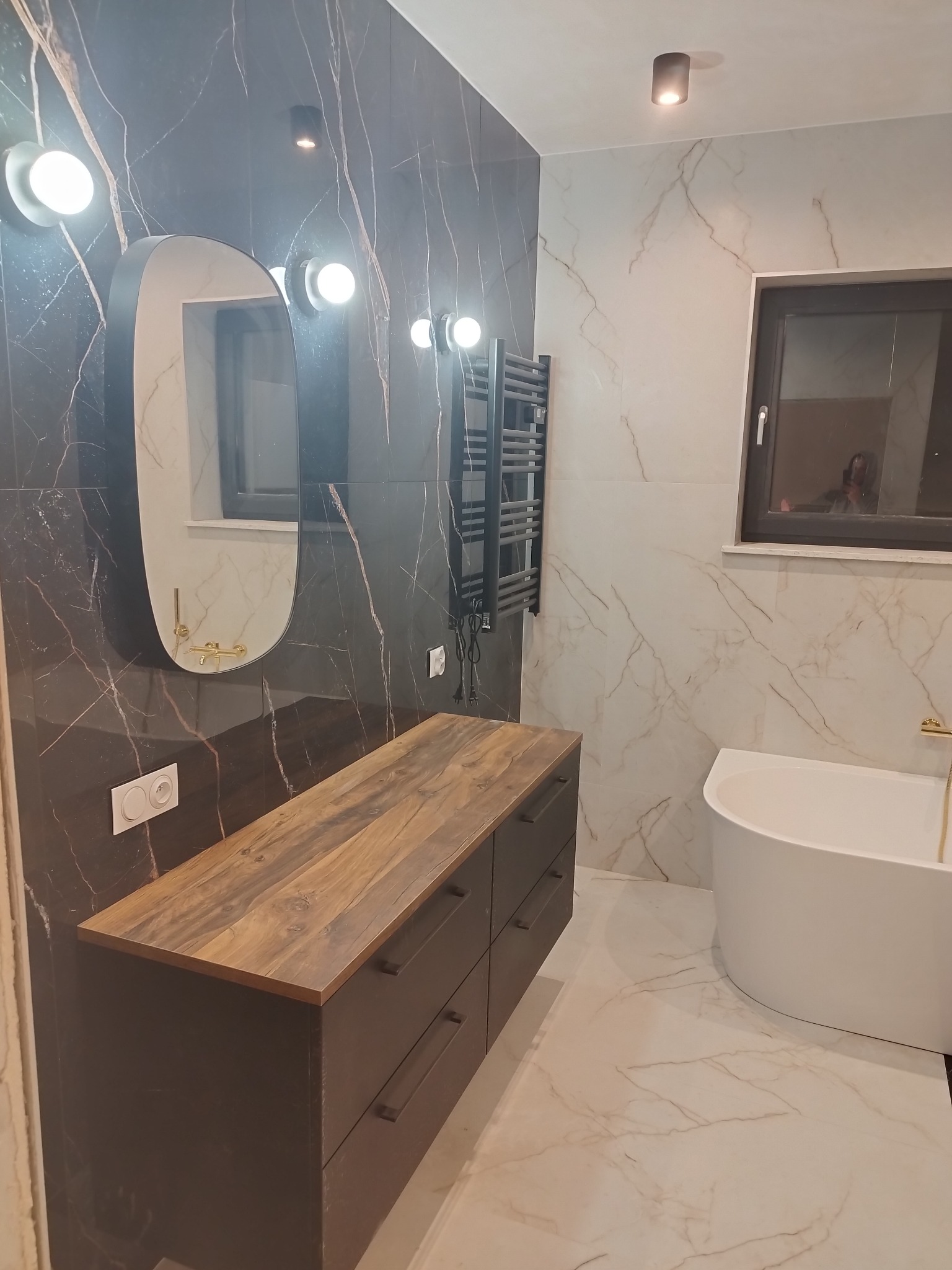 Nowoczesna łazienka z czarno-białymi marmurkowymi płytkami, owalnym lustrem z podświetleniem, drewnianą szafką pod umywalką i wolnostojącą wanną.