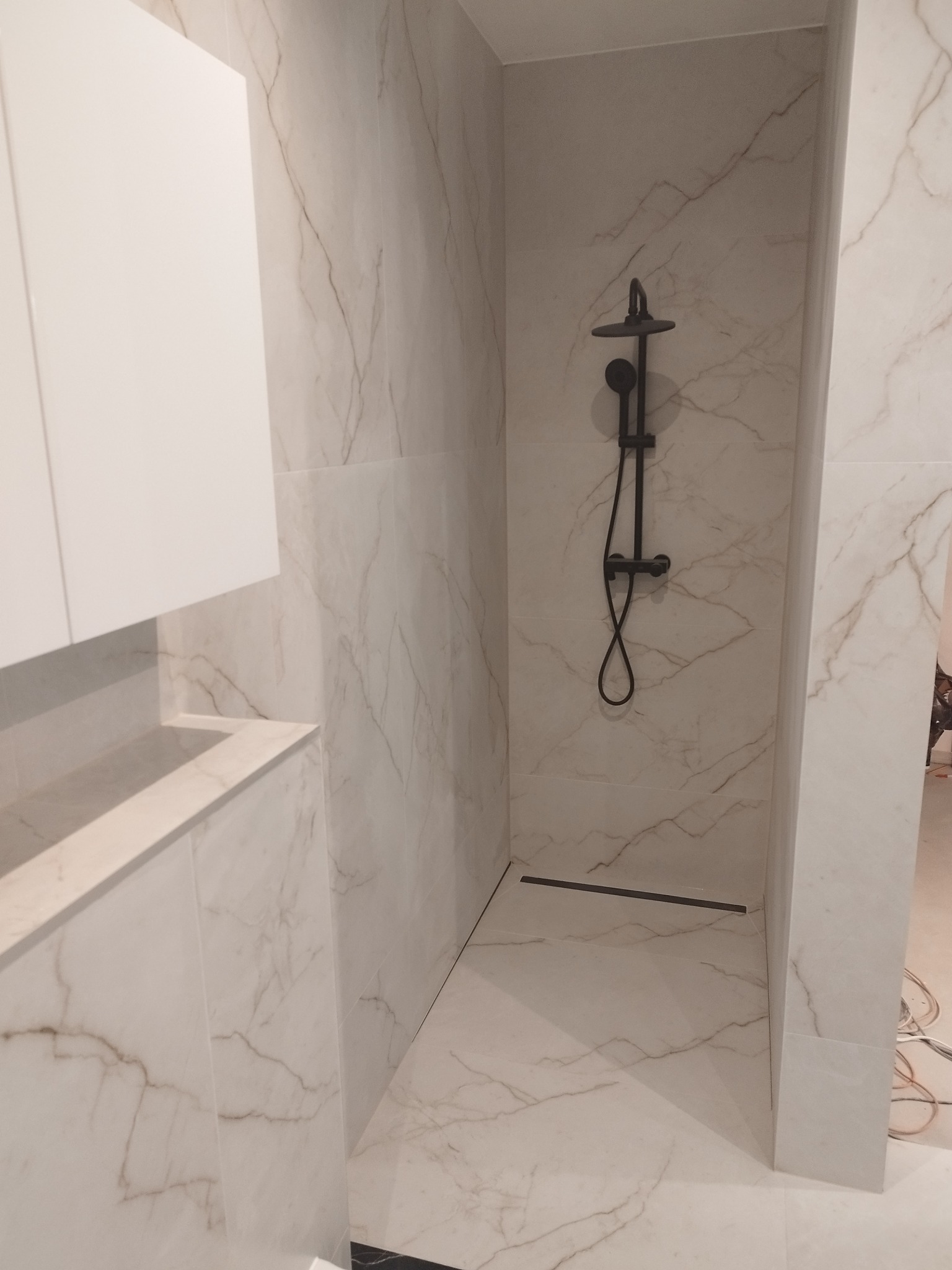 Kabina prysznicowa z czarną armaturą, wykończona płytkami imitującymi marmur, z odpływem liniowym w podłodze i szafką z umywalką po lewej stronie.