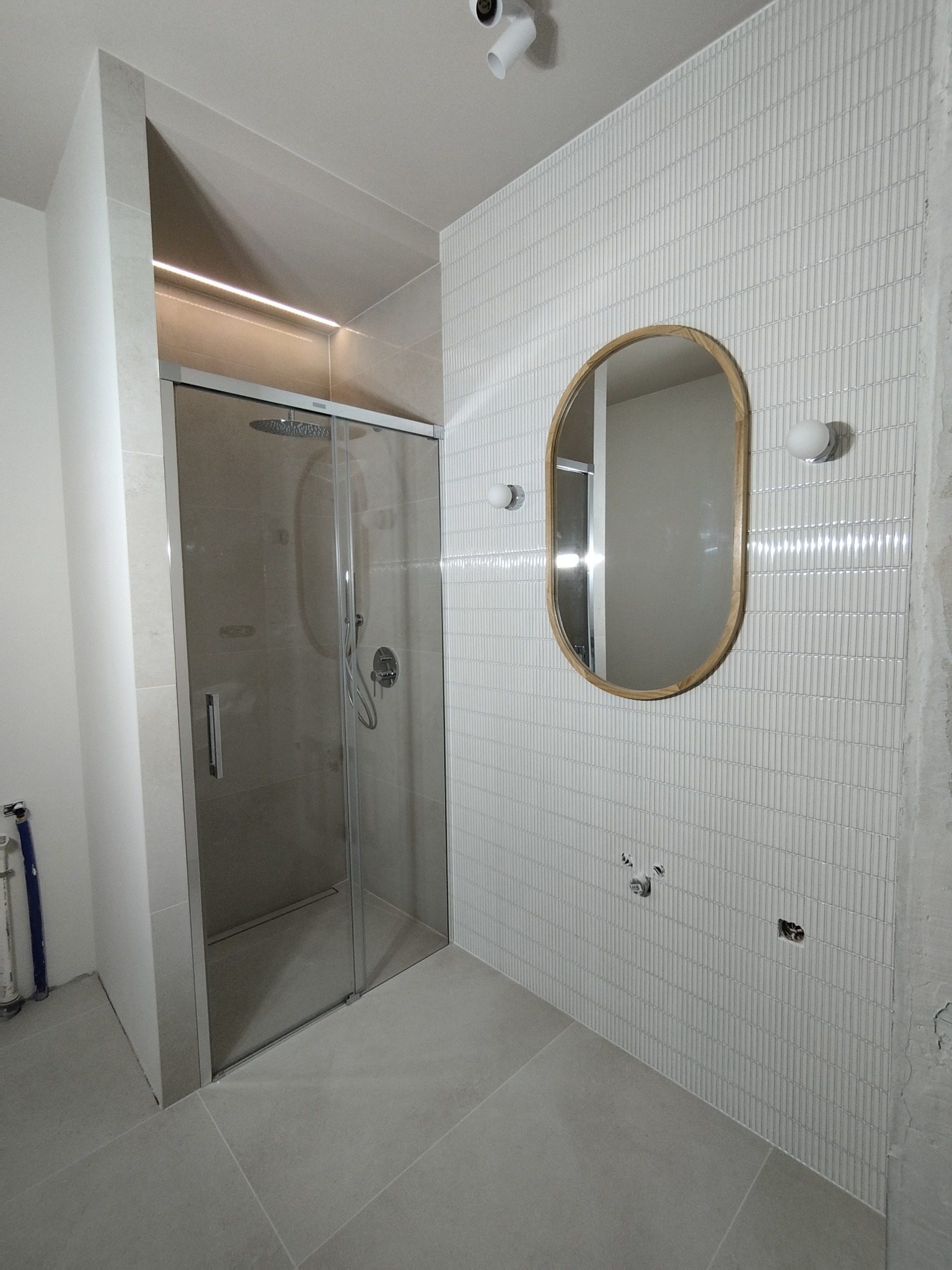 Wnętrze łazienki w trakcie wykańczania, widoczna kabina prysznicowa z deszczownicą, owalne lustro w drewnianej ramie, białe płytki ułożone pionowo oraz instalacja elektryczna w ścianie.