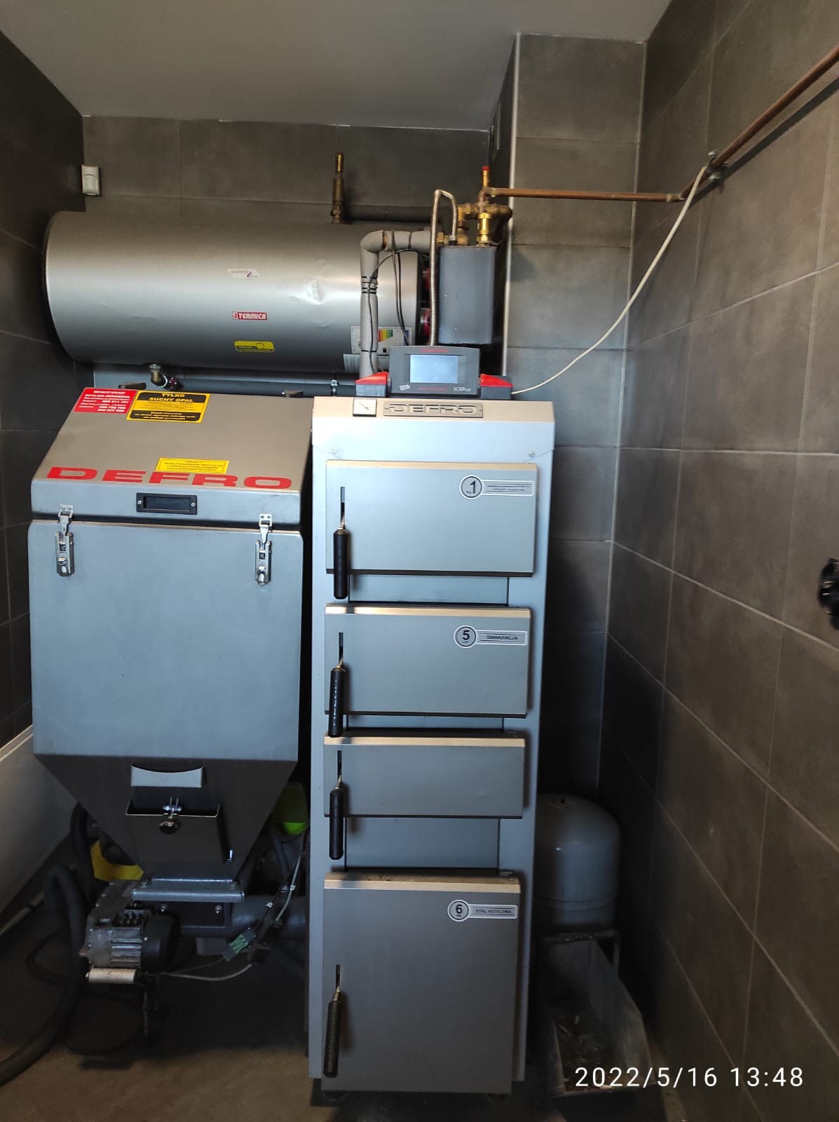 Szary piec CO marki Defro z podajnikiem, zbiornikiem na wodę i instalacją hydrauliczną na tle ciemnych płytek ściennych.