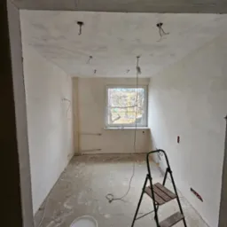 Wnętrze pokoju w trakcie malowania ścian na biało, widoczne ślady po szpachlowaniu, drabina malarska, wiadro z farbą, wystające kable oświetleniowe z sufitu.