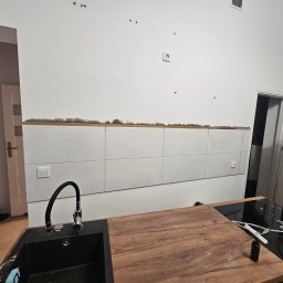 Kamil Mroczek - Kuchnia w trakcie prac wykończeniowych: częściowo ułożone białe płytki na ścianie, widoczne ślady po kleju, drewniany blat, czarny zlew z baterią, płyta indukcyjna z resztkami po remoncie, widoczne...