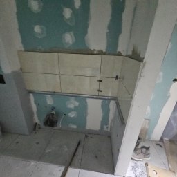 Kamil Mroczek - Ściana w trakcie układania płytek ceramicznych, widoczne krzyżyki dystansowe, surowy stan wykończenia ścian i podłogi, instalacja podtynkowa.