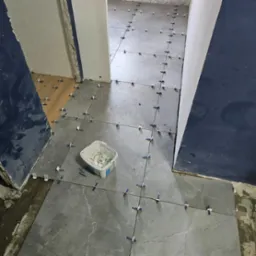 Układanie szarych płytek podłogowych w łazience, widoczne krzyżyki dystansowe i wiaderko z klejem, fragmenty drewnianej podłogi w sąsiednim pomieszczeniu.