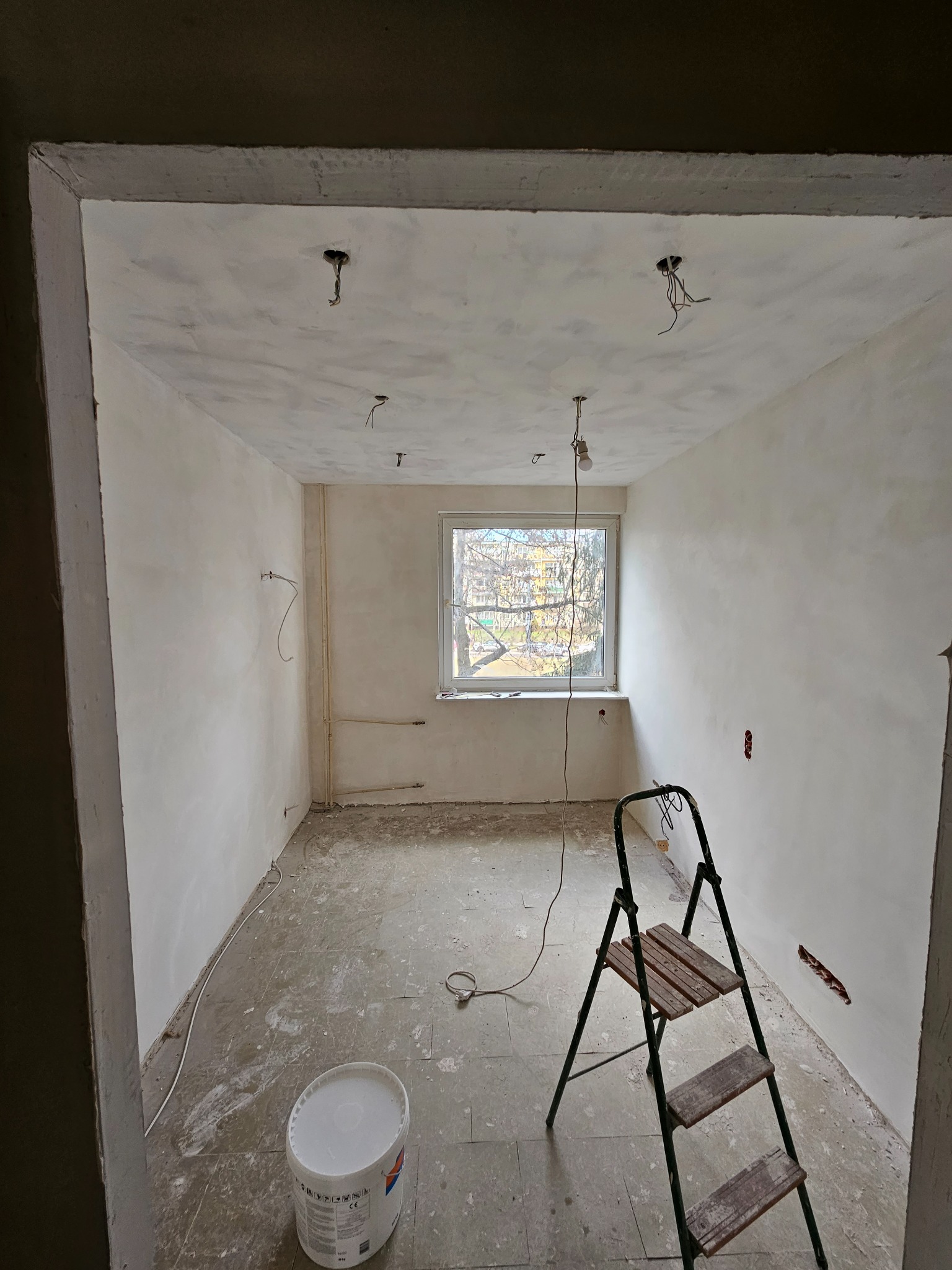 Wnętrze pokoju w trakcie malowania ścian na biało, widoczne ślady po szpachlowaniu, drabina malarska, wiadro z farbą, wystające kable oświetleniowe z sufitu.