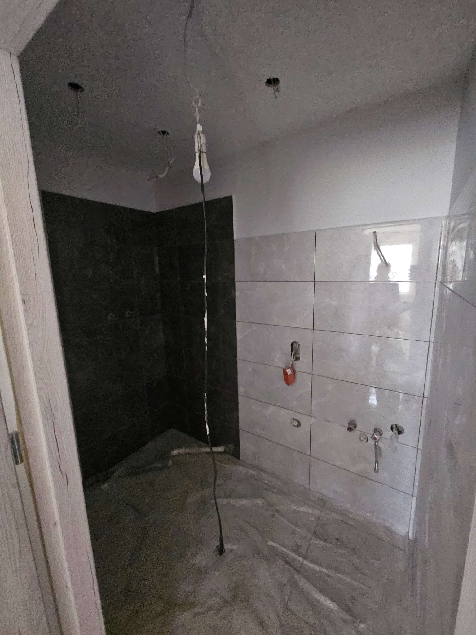 Wnętrze łazienki w trakcie prac wykończeniowych: częściowo ułożone płytki ścienne w jasnym odcieniu, ciemne płytki w strefie prysznica, surowa podłoga, widoczne instalacje wodne i prowizoryczne...