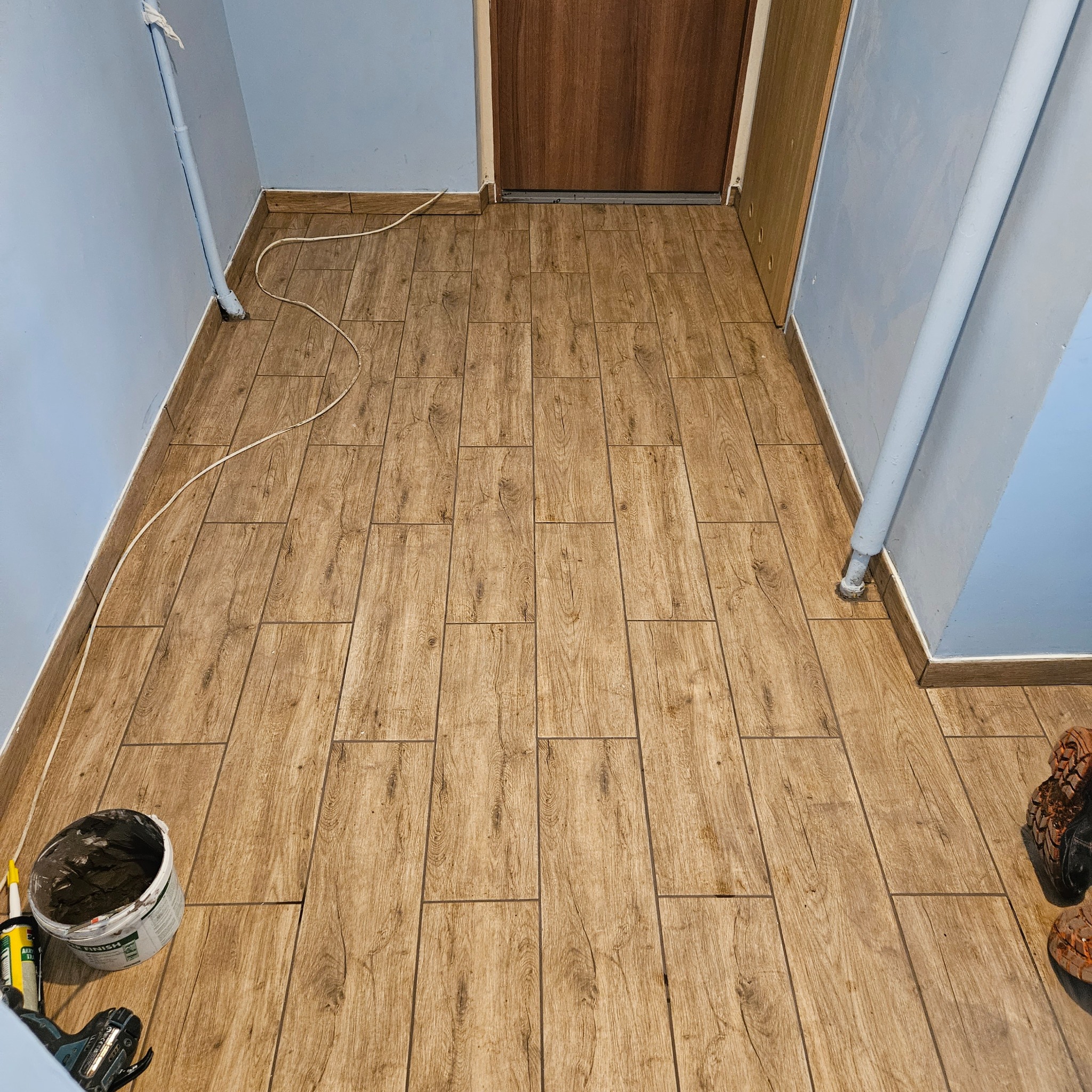 Ułożone panele podłogowe imitujące drewno w korytarzu, widoczne narzędzia i wiadro z klejem, częściowo ukończona praca.