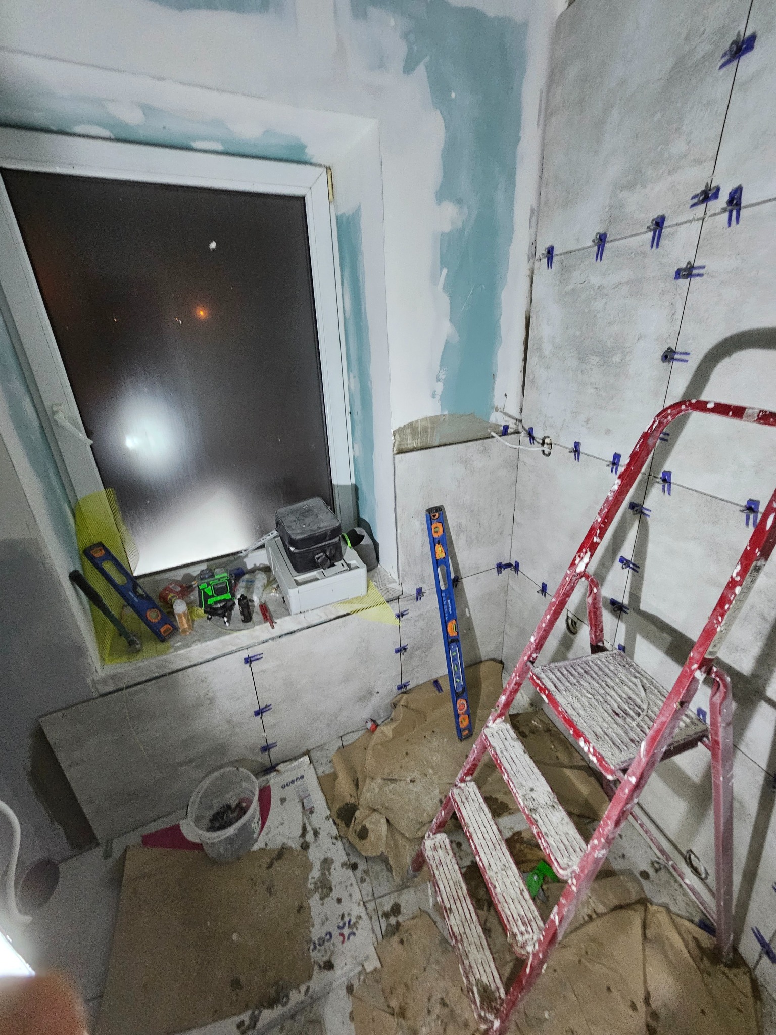 Wnętrze łazienki w trakcie remontu, widoczne płytki ścienne z systemem poziomowania, drabina malarska, okno z narzędziami i poziomice.