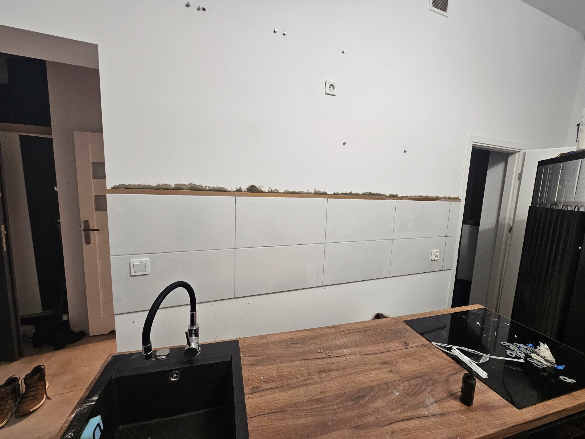 Kuchnia w trakcie prac wykończeniowych: częściowo ułożone białe płytki na ścianie, widoczne ślady po kleju, drewniany blat, czarny zlew z baterią, płyta indukcyjna z resztkami po remoncie, widoczne...