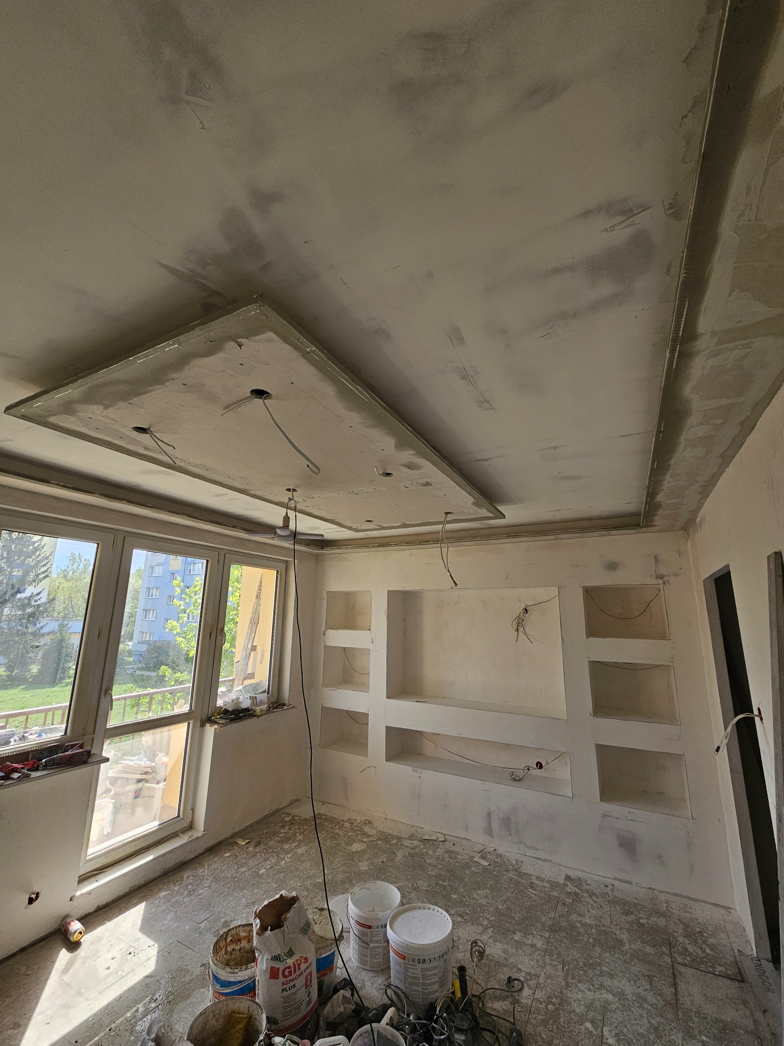 Wnętrze w trakcie wykańczania: surowy sufit z podwieszaną konstrukcją, ściany z wnękami, widoczne przewody elektryczne, podłoga zabezpieczona folią, wiadra z zaprawą gipsową i narzędzia malarskie.