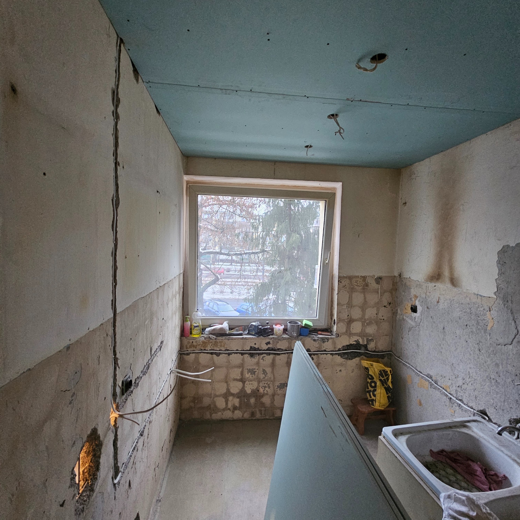 Kuchnia w trakcie remontu: odsłonięte przewody elektryczne, częściowo usunięte płytki, sufit pokryty płytą gipsową, okno z widokiem na zimowe drzewa, przygotowane narzędzia i materiały budowlane.