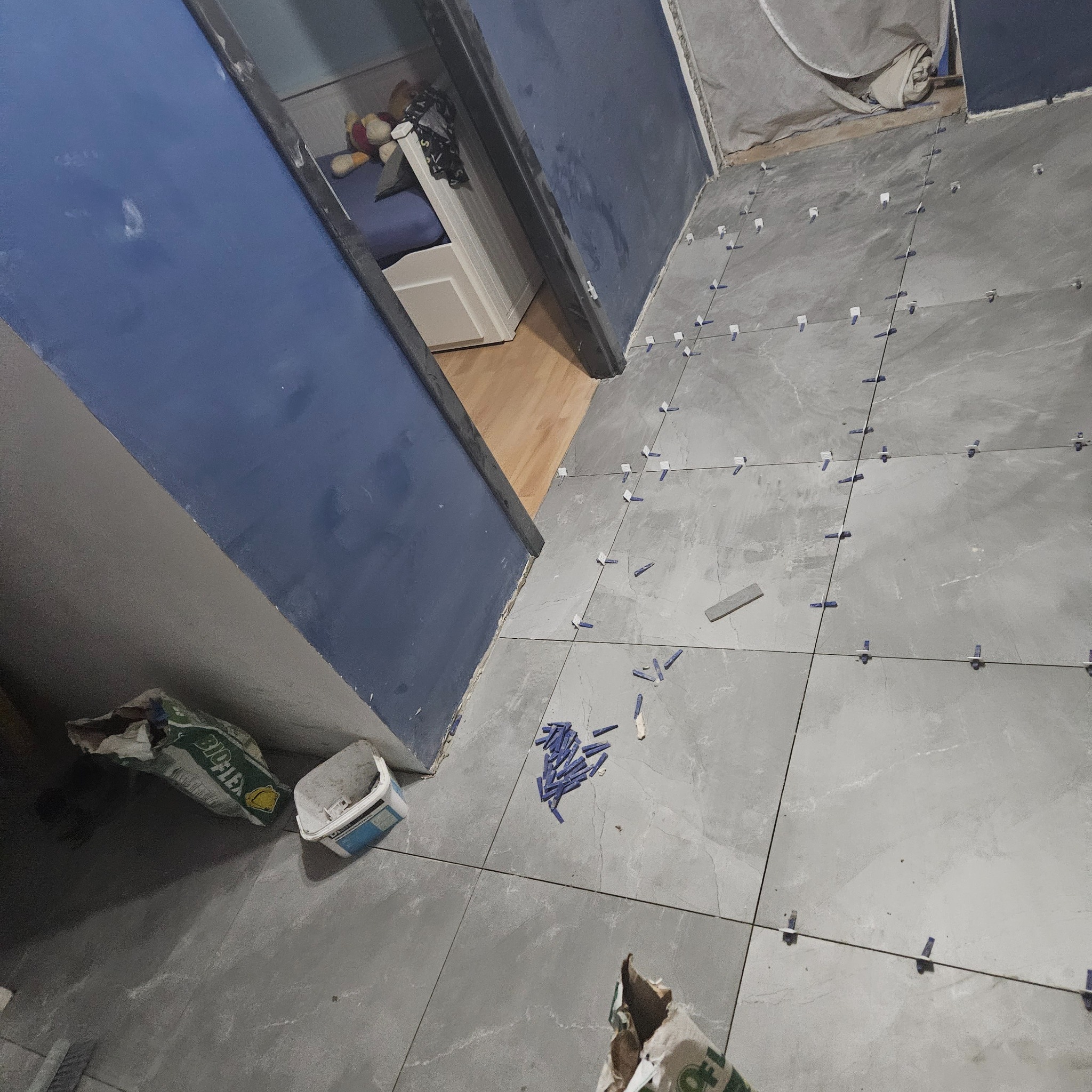 Szare płytki podłogowe ułożone z użyciem niebieskich krzyżyków dystansowych, widoczny fragment niebieskiej ściany i torba z klejem.