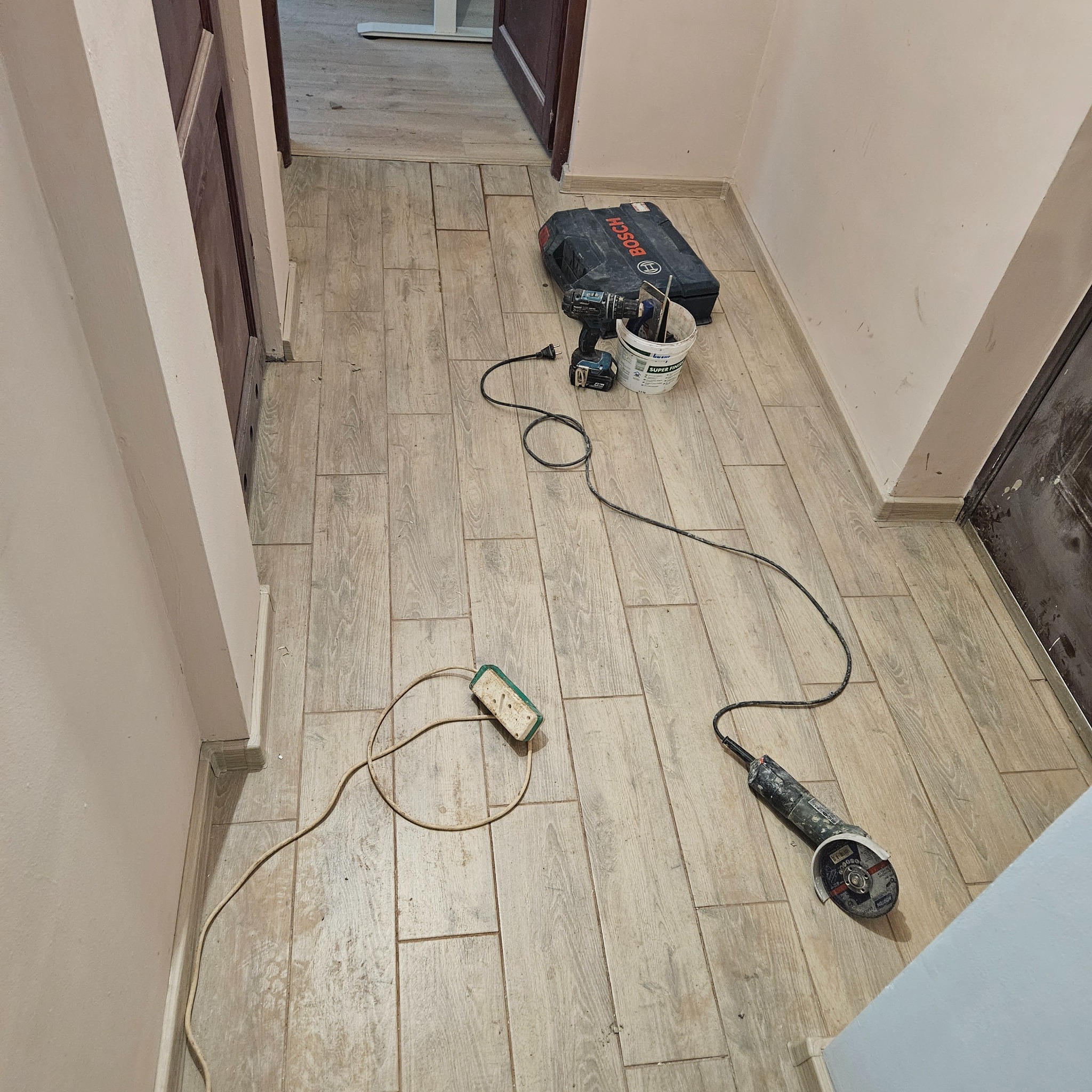 Ułożone panele podłogowe w korytarzu, widoczna szlifierka kątowa, przedłużacz, wiadro z narzędziami i skrzynka narzędziowa Bosch.