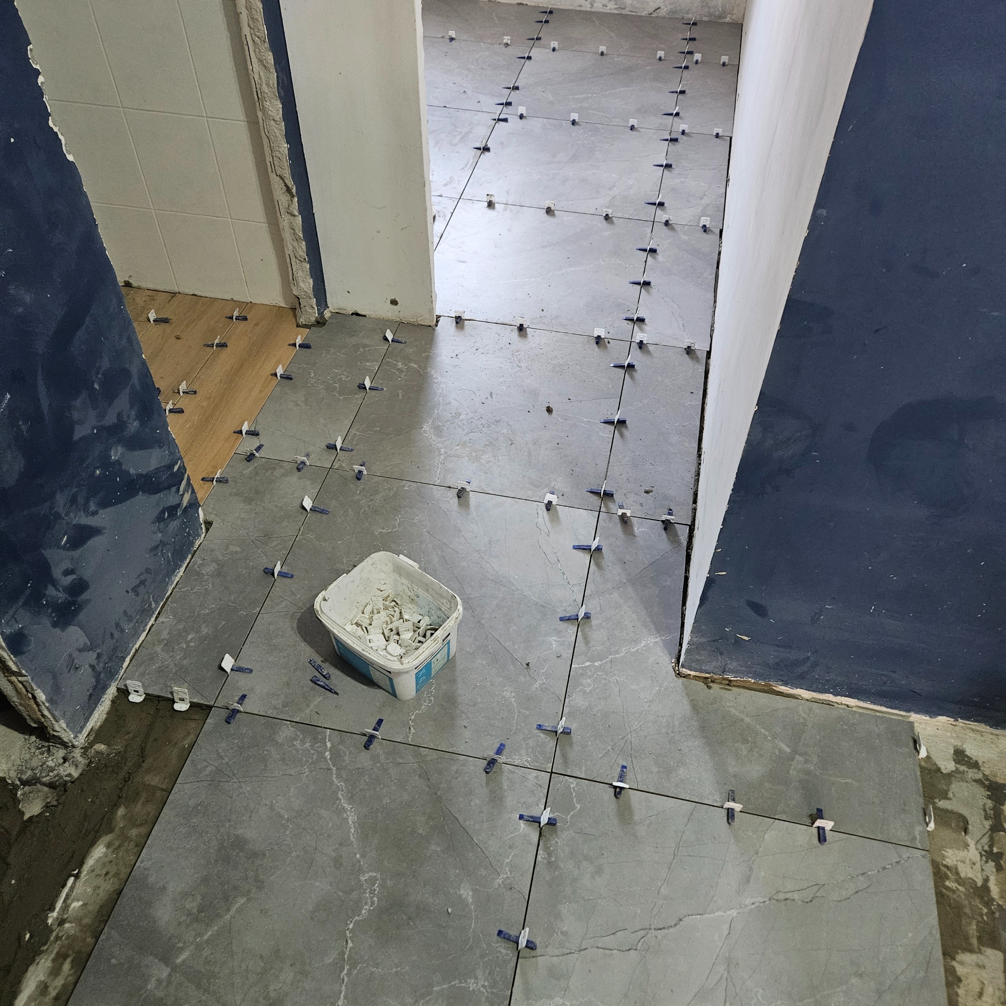 Układanie szarych płytek podłogowych w łazience, widoczne krzyżyki dystansowe i wiaderko z klejem, fragmenty drewnianej podłogi w sąsiednim pomieszczeniu.