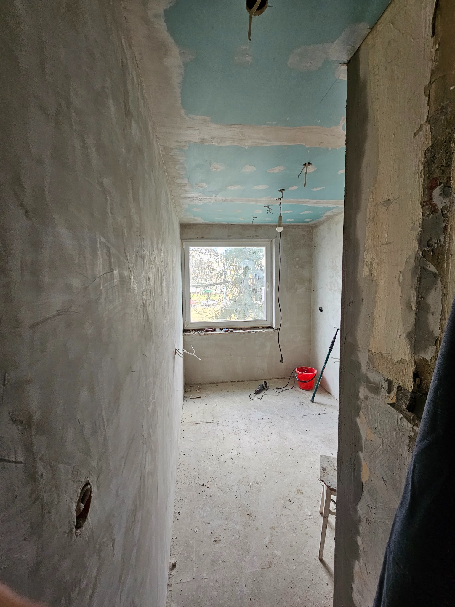 Pokój w trakcie prac wykończeniowych: surowe ściany pokryte gładzią, fragment sufitu z pozostałościami niebieskiej farby, prowizoryczne oświetlenie, czerwone wiadro i szlifierka kątowa przy oknie...