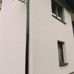Krzysztof Stępień - Fragment elewacji budynku po nałożeniu tynku strukturalnego w odcieniach beżu, widoczna brązowa rynna spustowa i dwa okna z brązowymi ramami, na ścianie zamontowana antena satelitarna.