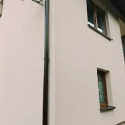 Fragment elewacji budynku po nałożeniu tynku strukturalnego w odcieniach beżu, widoczna brązowa rynna spustowa i dwa okna z brązowymi ramami, na ścianie zamontowana antena satelitarna.