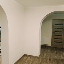 KrisBud - Wnętrze po remoncie: łukowate przejście, beżowe ściany, drewnopodobne płytki podłogowe, widoczny fragment drzwi i grzejnika.