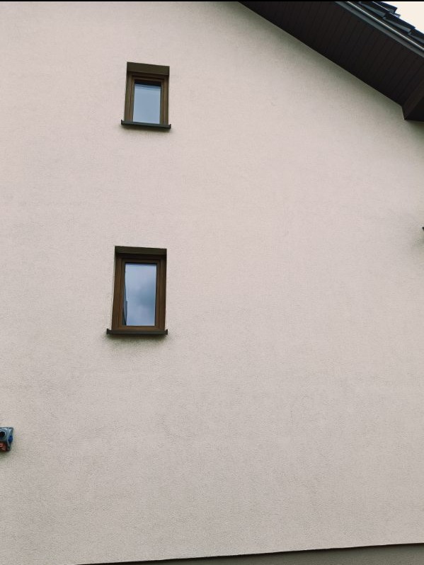 Fragment elewacji budynku z dwoma małymi oknami o brązowych ramach, widoczna faktura tynku baranek i fragment dachu z ciemną podbitką.