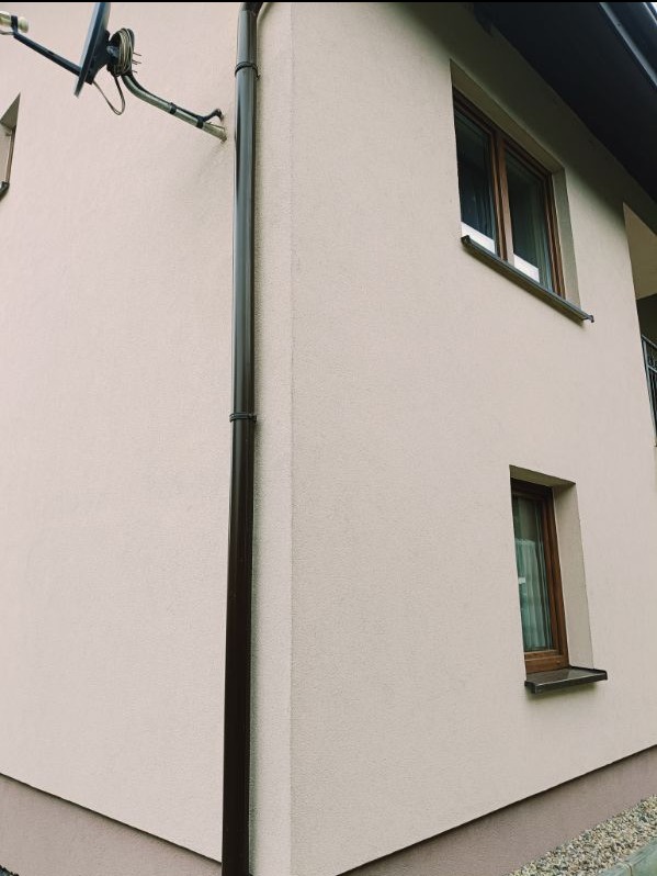 Fragment elewacji budynku po nałożeniu tynku strukturalnego w odcieniach beżu, widoczna brązowa rynna spustowa i dwa okna z brązowymi ramami, na ścianie zamontowana antena satelitarna.