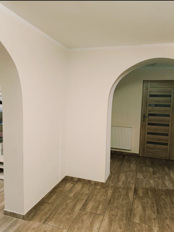 Wnętrze po remoncie: łukowate przejście, beżowe ściany, drewnopodobne płytki podłogowe, widoczny fragment drzwi i grzejnika.