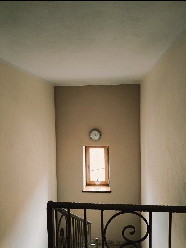 Klatka schodowa z beżowymi ścianami, drewnianym oknem, zegarem na ścianie i czarną, metalową barierką.