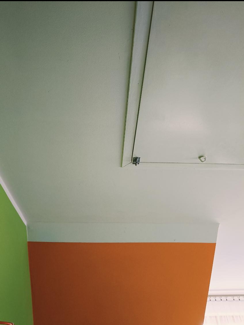 Narożnik pokoju z pomalowanymi ścianami w kolorach zielonym i pomarańczowym, biały sufit z klapą rewizyjną i widocznym zamkiem.