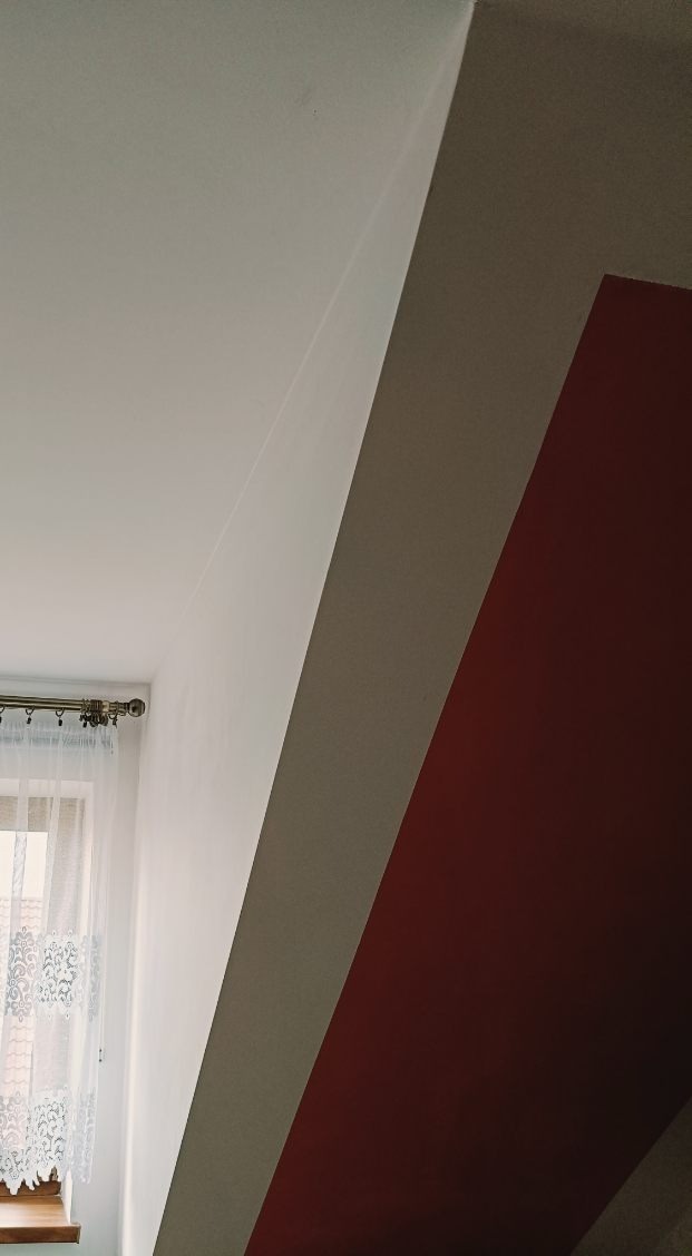 Pomieszczenie po malowaniu z widoczną białą ścianą, fragmentem okna z firanką i skośną ścianą pomalowaną na bordowo.