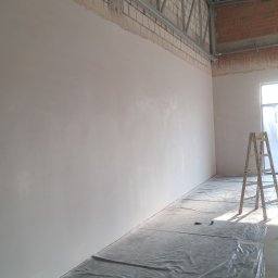 Usługi Remontowo Wykończeniowe Kacper Jambor - Świeżo pomalowana, jasna ściana w surowym wnętrzu z widoczną konstrukcją stropu i ceglanymi fragmentami. Podłoga zabezpieczona folią malarską, w tle drabina.