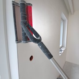 Usługi Remontowo Wykończeniowe Kacper Jambor - Szlifowanie ściany szlifierką z długim uchwytem, widoczny otwór na gniazdko elektryczne i okno z narzędziami na parapecie.