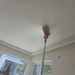 Usługi Remontowo Wykończeniowe Kacper Jambor - Szlifowanie sufitu za pomocą szlifierki na wysięgniku w pomieszczeniu z oknami i listwami dekoracyjnymi.