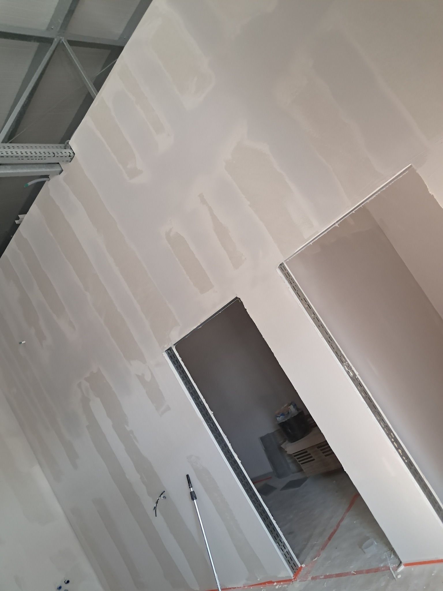 Ściana po nałożeniu pierwszej warstwy gładzi, widoczne ślady po szpachli, dwa otwory drzwiowe z metalowymi profilami, w tle pomieszczenie w trakcie prac wykończeniowych.